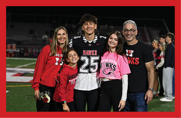 Four people standing on a sports field wearing athletic clothing and cleats with one person holding a bouquet of flowers. Description generated by AI