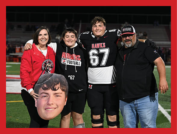 Four people stand on a football field at night with one person wearing a football uniform and another holding a large cutout of a smiling face. Description generated by AI