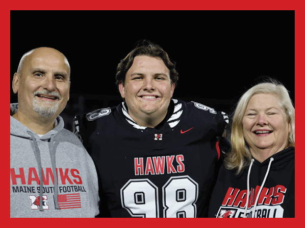 A person in a football uniform with the number 74 stands between two adults on a football field at night, with one adult holding a flower. Description generated by AI