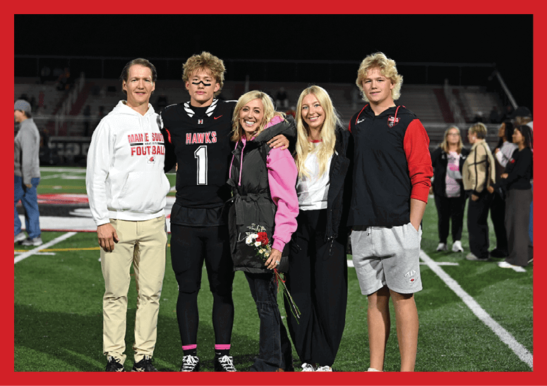 Five people stand closely together on a sports field at night with one person holding flowers and others wearing casual and sports clothing. Description generated by AI