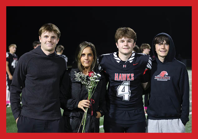 Three people stand side by side on a field, with one person holding a bouquet of flowers and another wearing a sports jersey with the word “HAWKS" and the number 4. Description generated by AI