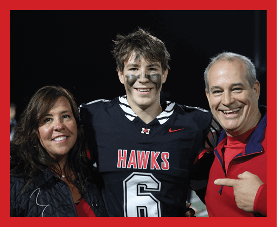 Three people stand closely together smiling, with one wearing a black football jersey labeled “HAWKS" and the number 6, while the others wear casual jackets. Description generated by AI