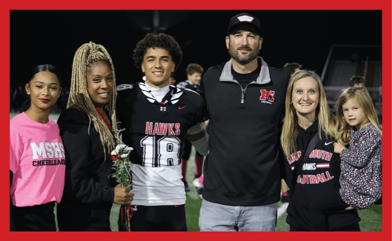 Four people stand together on a sports field at night, with one person holding flowers, another wearing a football uniform, and another holding a child dressed in a patterned outfit. Description generated by AI