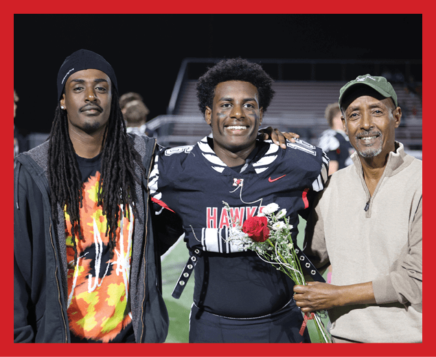 Two adults stand side by side on a sports field at night, one wearing a colorful shirt and jacket and the other in a football uniform holding a bouquet of flowers and smiling. Description generated by AI