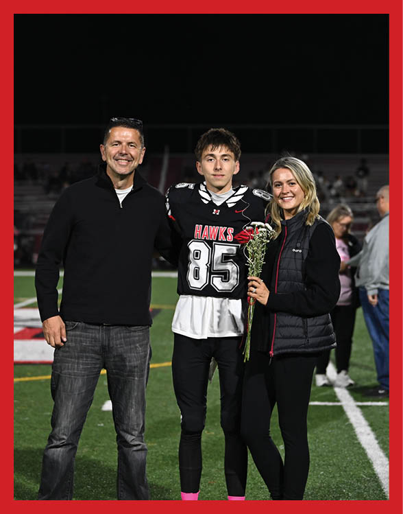 Three people stand together on a sports field with one person in a football uniform holding flowers and the others dressed casually. Description generated by AI