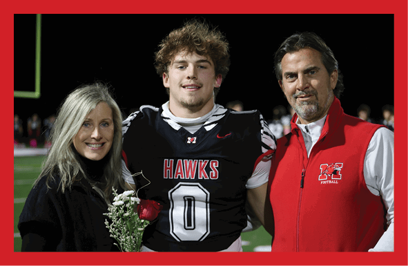 Three people stand together on a sports field with one person in a black football jersey holding a red rose and white flowers, flanked by others wearing black and red jackets. Description generated by AI