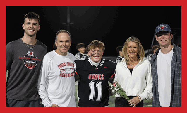 Five people stand closely together on a sports field at night with one person in a football uniform holding flowers and smiling. Description generated by AI