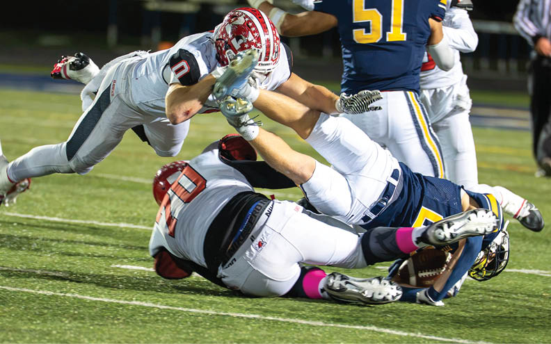 A person in a white football uniform with the number 10 is diving forward while another person in a red helmet and white uniform is on the ground attempting to tackle them during a game on a grassy field. Description generated by AI