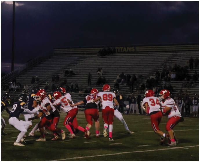 People wearing football uniforms are playing a game on a field under stadium lights with a dark cloudy sky in the background. Description generated by AI