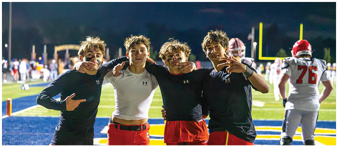 Four people wearing athletic clothing pose together on a sports field at night with a football goalpost and other players visible in the background. Description generated by AI