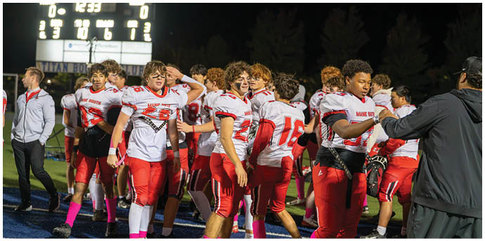 A group of people wearing red and white football uniforms are gathered on a field at night near a scoreboard. Description generated by AI