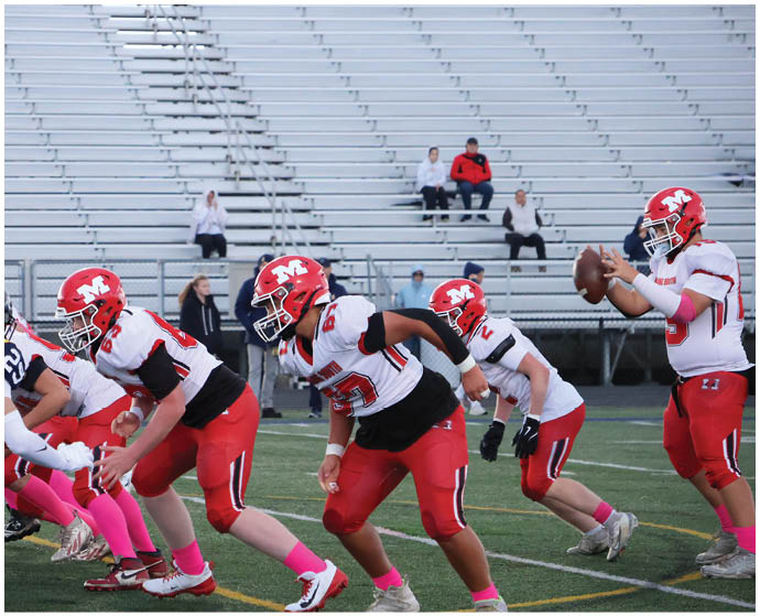 Adults in football uniforms with red helmets and pink socks are lined up on a field facing opponents in dark uniforms during a game. Description generated by AI