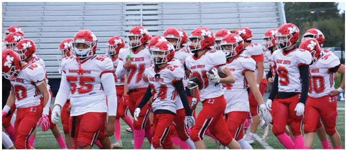 A group of people wearing red and white football uniforms and helmets are gathered on a field with empty bleachers in the background. Description generated by AI
