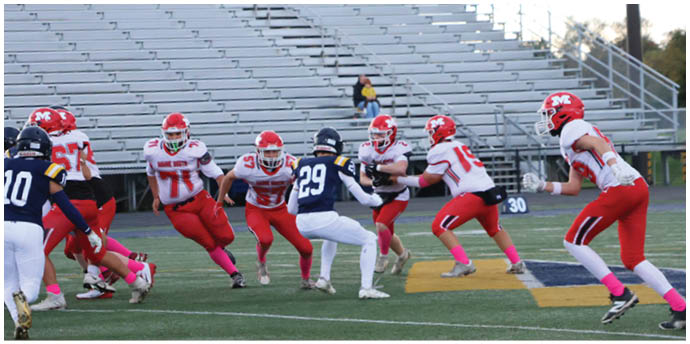 People wearing red and white football uniforms are running and preparing to block players in dark blue and white uniforms on a football field with empty bleachers in the background. Description generated by AI
