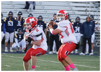 Two people in red and white football uniforms are on a field with one preparing to throw a football and the other running, while a group of people in dark uniforms stand on the sidelines in front of empty bleachers. Description generated by AI