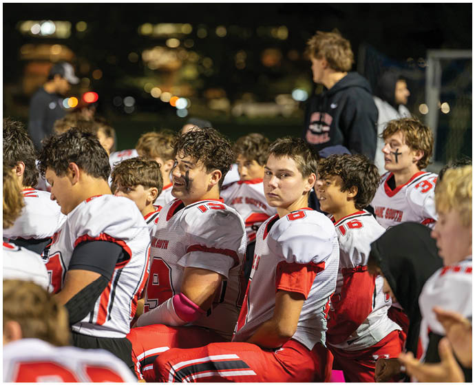 A group of people wearing white and red football uniforms are gathered closely together, some sitting and some standing, with one person smiling and others looking attentive. Description generated by AI