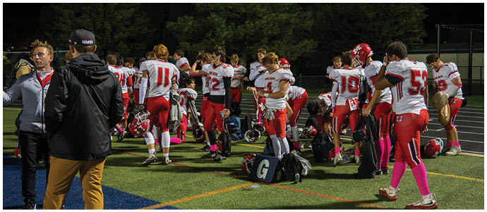 People in red and white football uniforms are gathered on a field, some standing and some sitting, with two people in casual clothing in the foreground. Description generated by AI