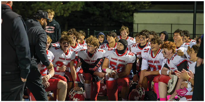 A group of people wearing red and white football uniforms are sitting and kneeling on the sidelines of a field, some holding helmets and others clapping. Description generated by AI