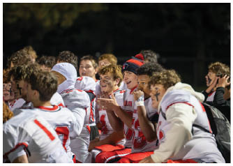 A group of people in sports uniforms with face paint are sitting closely together, some clapping and smiling, likely on the sidelines of a game. Description generated by AI