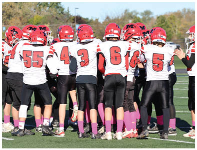 People in football uniforms with red helmets and pink socks are lined up on a field facing each other in a ready stance under a clear blue sky. Description generated by AI