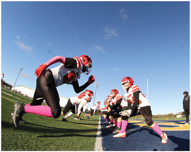A group of people wearing red helmets, white jerseys with large numbers, black pants, and pink socks are standing together on a sports field. Description generated by AI