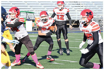Children playing a football game on a field with one player being tackled while others approach, and a group of players and coaches stand on the sidelines near empty bleachers. Description generated by AI