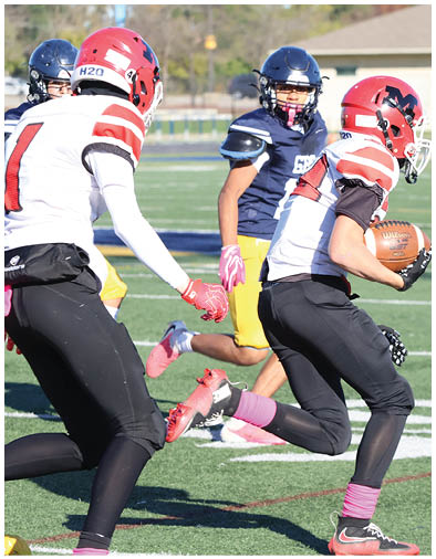 Two people in football uniforms are running on a field with one holding a football while others in the background appear to be chasing them. Description generated by AI