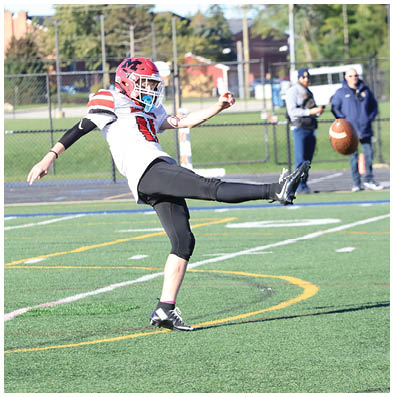A person wearing a red helmet and white sports jersey is kicking a football on a green field with two people standing in the background near a fence. Description generated by AI