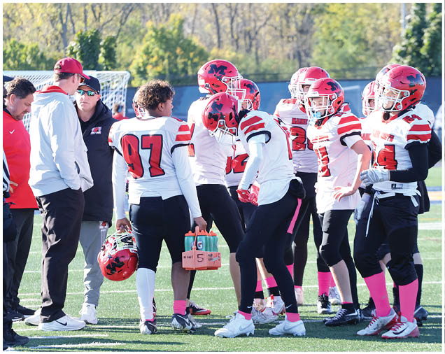 A group of people wearing red helmets, white jerseys with red and black stripes, black pants, and pink socks are standing together on a sports field, with one person holding a carrier of water bottles. Description generated by AI