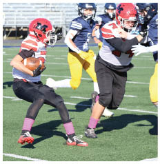 Two people in football uniforms with red helmets and pink socks are playing on a field while a third person in a navy and yellow uniform runs behind them. Description generated by AI