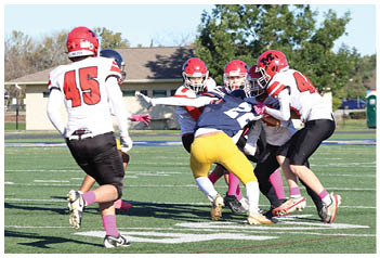 Several people wearing football helmets and uniforms are engaged in a play on a grassy field with a building and trees in the background. Description generated by AI