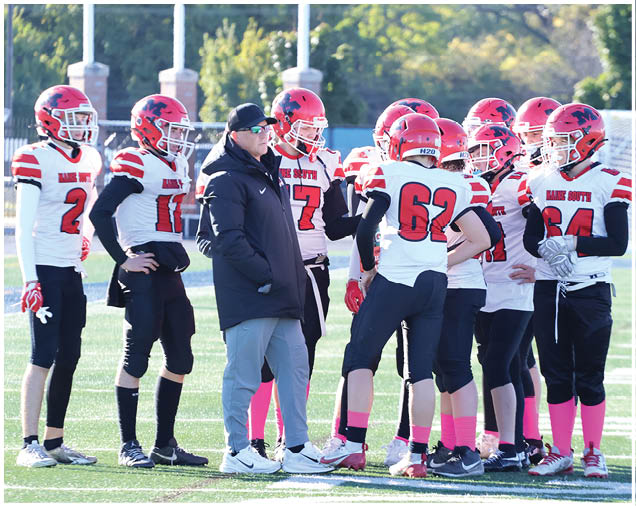A group of people wearing red helmets and white sports jerseys with black pants are gathered on a football field with one person in a black jacket and gray pants standing among them. Description generated by AI