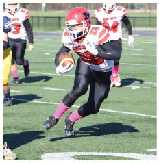 A person wearing a red helmet and black and white football uniform is running with a football on a green field while other players in various uniforms pursue. Description generated by AI