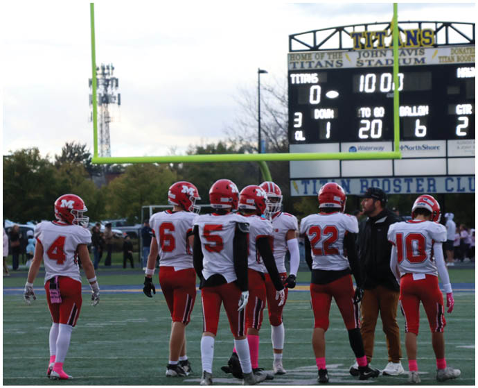 A group of people wearing red and white football uniforms are standing on a field near a goalpost with a scoreboard in the background showing a score and game details. Description generated by AI