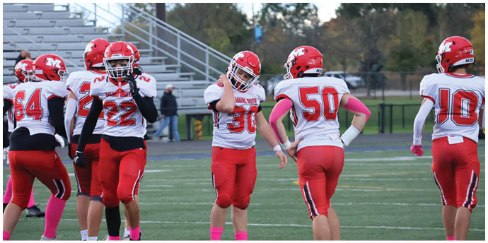 A group of people wearing red and white football uniforms are standing on a field with bleachers and trees in the background. Description generated by AI