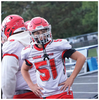 A person wearing a red football helmet and white jersey with the number 51 stands with hands on hips on a sports field. Description generated by AI