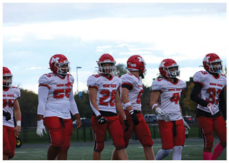 Four people wearing red and white football uniforms and helmets are standing on a field under a cloudy sky. Description generated by AI