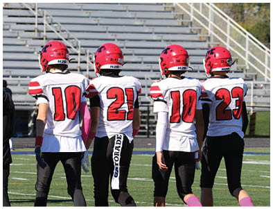 Four people wearing red helmets and white football jerseys with numbers stand on a sports field facing away from the camera with empty bleachers in the background. Description generated by AI