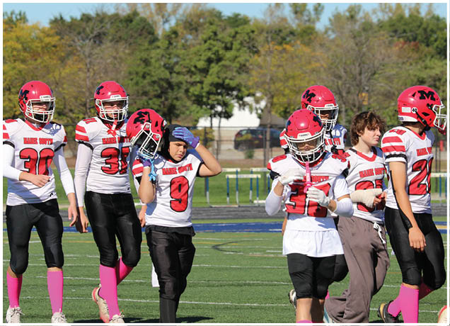 Five people wearing red helmets and white football jerseys with black pants are standing on a football field with trees and a fence in the background. Description generated by AI