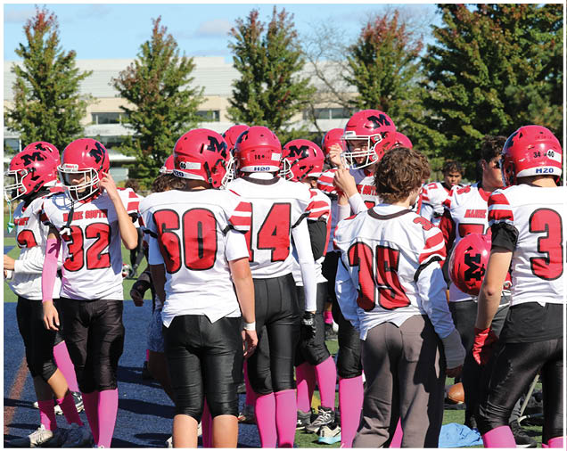 A group of people wearing red helmets, white and red sports jerseys, black pants, and pink socks are gathered on a sports field with trees and a building in the background. Description generated by AI