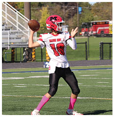 A person wearing a red and white football uniform with the number 10 is preparing to throw a football on a grassy field with a fire truck and bleachers in the background. Description generated by AI