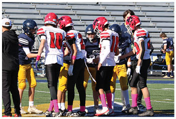 Two children on a football field are engaged in a game, with one running in a white and red uniform and the other kneeling while holding a football in a red sweatshirt and camouflage pants. Description generated by AI