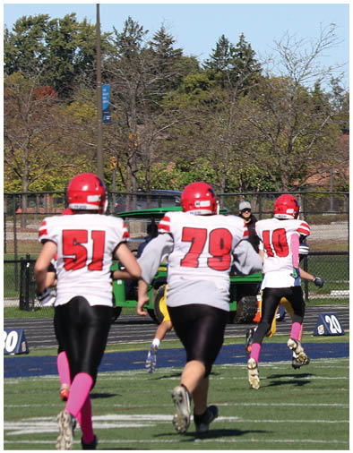 Several people wearing football helmets and jerseys are standing on a field with trees and houses in the background. Description generated by AI
