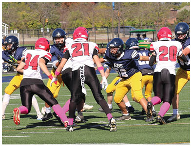 Several people wearing football uniforms in blue and yellow or white and red are actively engaged in a play on a green football field with trees and buildings in the background. Description generated by AI