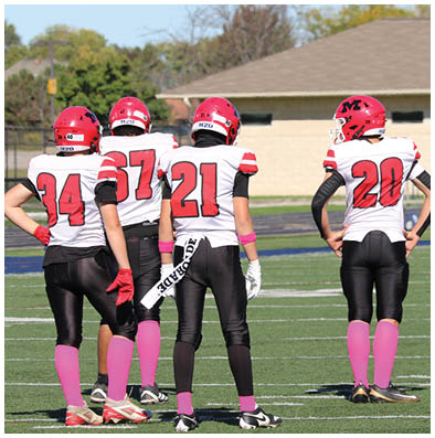 Two people in white and red football uniforms stand on a field near a football while a group of people in blue and yellow uniforms stand together in the background. Description generated by AI