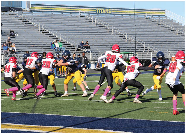A group of people wearing football uniforms in red and white or navy and yellow are actively playing on a football field with empty bleachers in the background. Description generated by AI