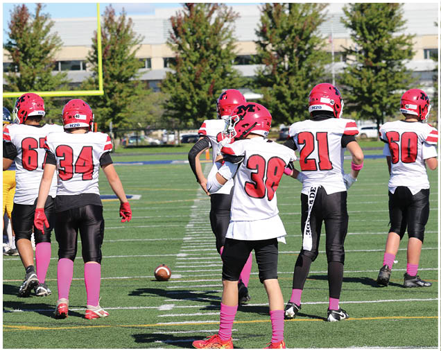 Children wearing red helmets, white jerseys with red numbers, black pants, and pink socks are standing on a football field near a football on the ground. Description generated by AI