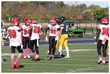 Adults wearing football uniforms in red, white, black, yellow, and blue are standing on a football field with trees and a green utility vehicle in the background. Description generated by AI