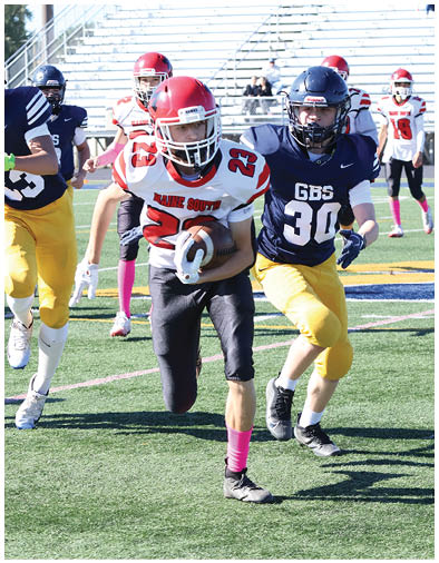 A person wearing a red helmet and white jersey with the number 23 is running with a football while being chased by people in blue jerseys and yellow pants on a sports field. Description generated by AI