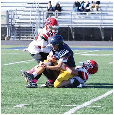 Three people wearing football helmets and uniforms are on a field with two tackling the third who is holding a football. Description generated by AI